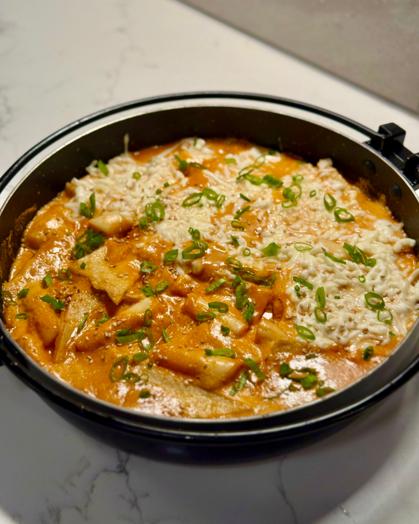 Close-up of rose tteokbokki in a skillet topped with melted mozzarella and chopped green onions.