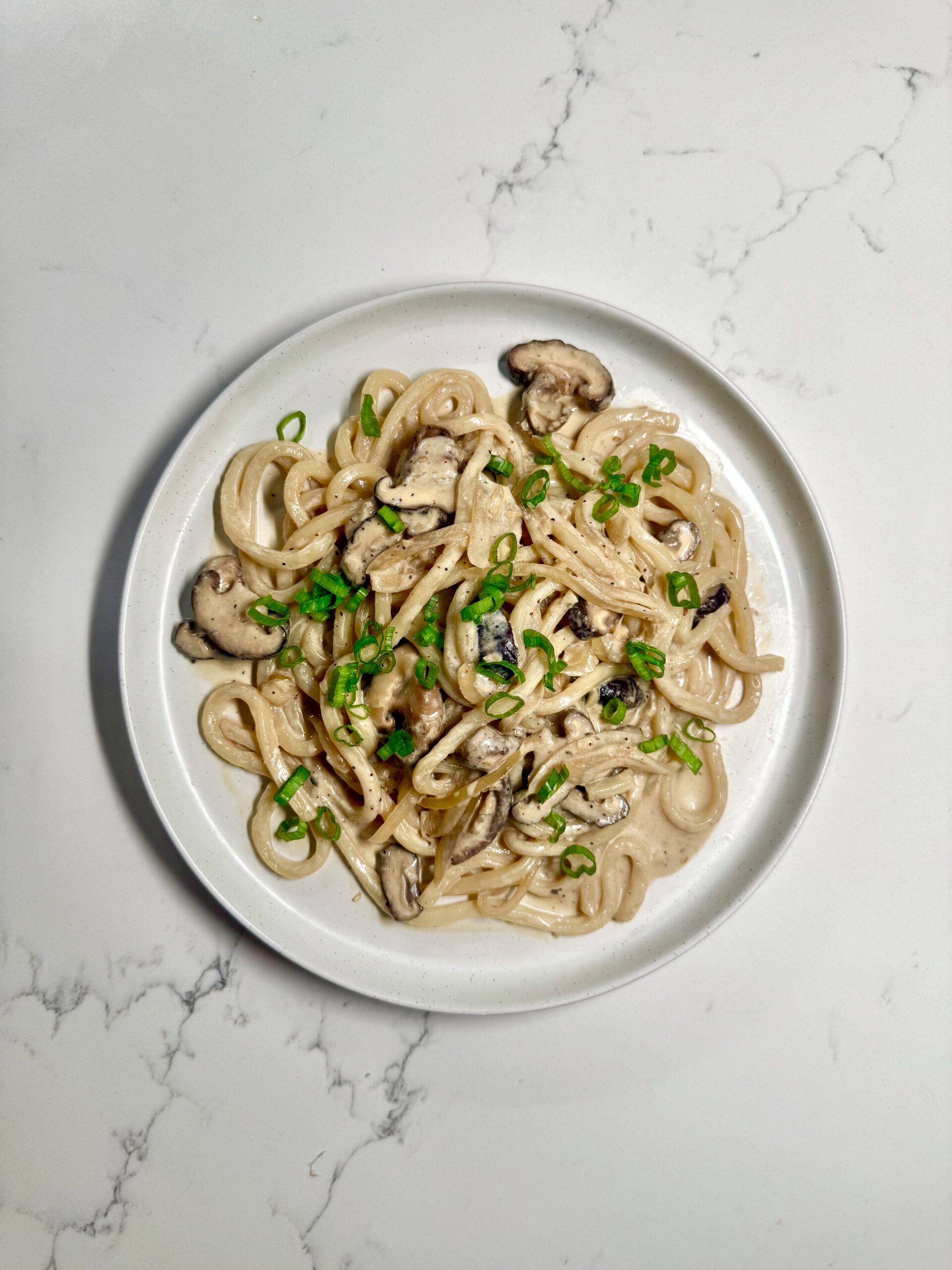 A bowl of creamy mushroom udon topped with sliced green onions and sesame seeds on a white countertop.