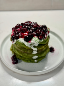 A tall stack of fluffy matcha pancakes topped with warm blueberry compote and fresh berries on a white plate.