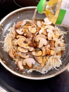 Sliced mushrooms being added to a sauté pan with onions and garlic.
