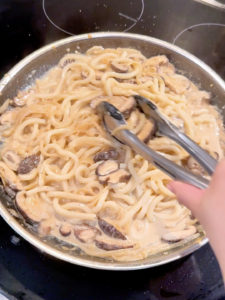 Udon noodles simmering in a creamy mushroom sauce in a pan on the stove.