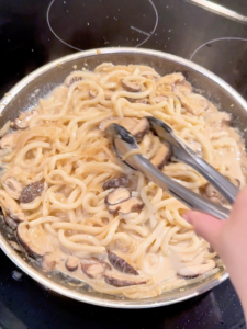 Udon noodles simmering in a creamy mushroom sauce in a pan on the stove.