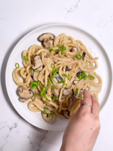 A hand reaching toward a bowl of creamy mushroom udon on a white surface.