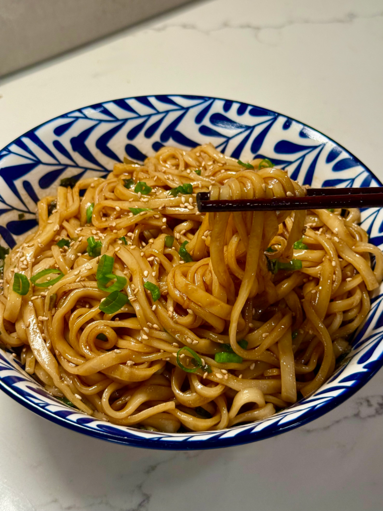 Close-up of scallion oil noodles being lifted with chopsticks, showing glossy soy-coated strands garnished with sesame seeds and green onions.