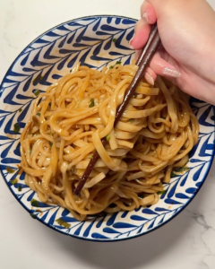 Hand mixing scallion oil noodles in a patterned bowl with chopsticks, coating noodles evenly in fragrant soy-scallion oil sauce.